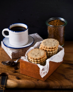 Two stacks of Coffee Shortbread Cookies in a small box. A cup of espresso and some coffee granules in the background.