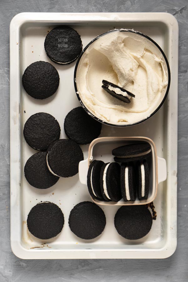 A batch of oreo cookies and a bowl of frosting on a baking tray.