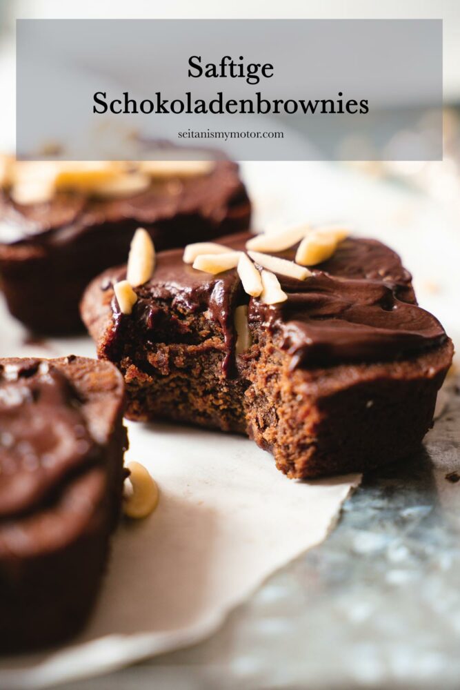 Das Bild zeigt saftige Schokoladenbrownies auf einer Unterlage aus Backpapier. Alle Brownies sind mit Ganache und Mandeln dekoriert, ein Brownie ist angebissen.