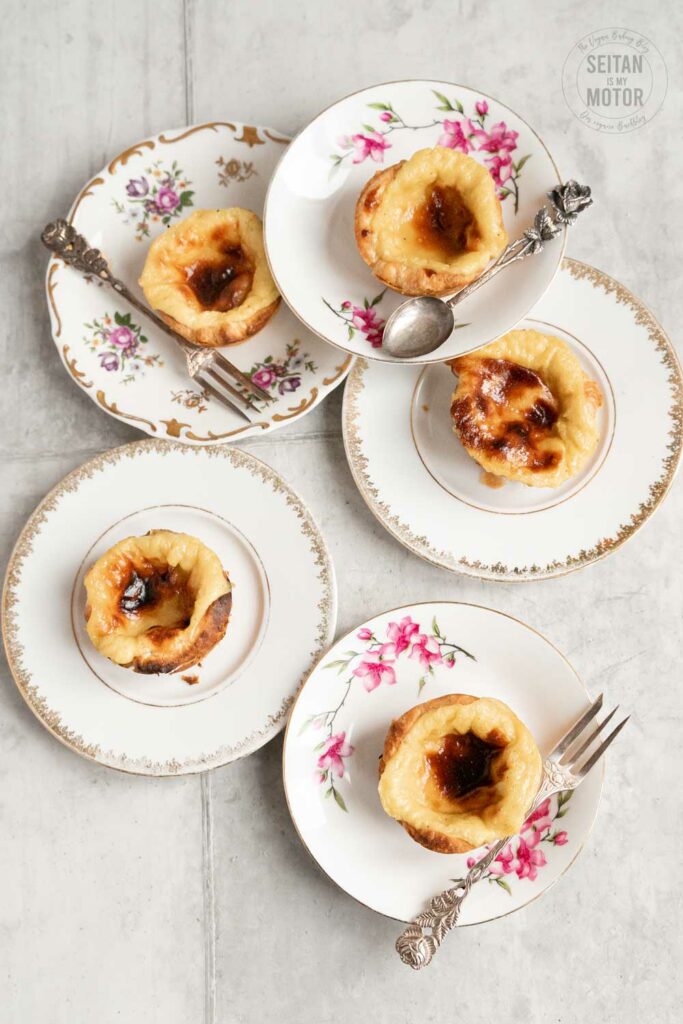 Five vegan pastéis de nata stiing on five decorative plates.